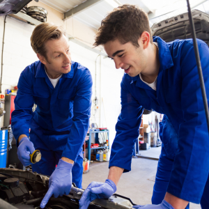 A mechanic wearing blue coveralls and latex gloves holds a piece of a car engine while pointing to an area under the hood while a younger mechanic apprentice watches and listens intently.