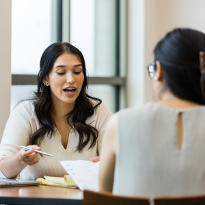 A woman with long dark hair sits across from another woman with dark hair and glasses showing and explaining documents she is handing to her.