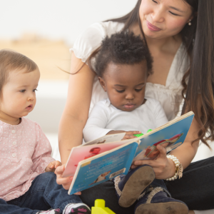 A young woman holds a baby in her lap while sitting on the floor with another baby sitting next to them looking at a book the woman has open for both babies to see.
